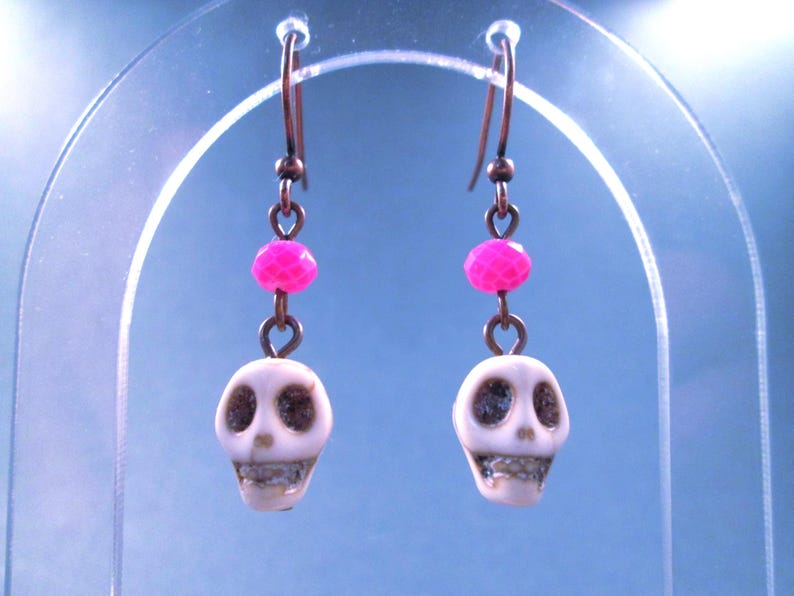 May include: A pair of drop earrings featuring white skull-shaped beads with dark eye sockets and teeth, accented by bright pink faceted beads. The skulls are suspended from copper-toned metal ear wires, creating a distinct contrast between the light skull beads and the vibrant pink accents.
