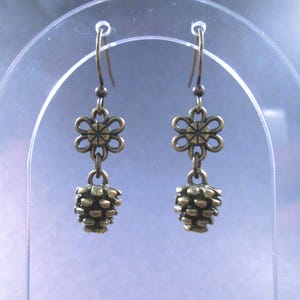 May include: A pair of bronze-toned dangle earrings featuring a small flower-shaped charm connected to a textured pinecone-shaped charm. The earrings hang from simple metal hooks and have a dark, metallic finish with visible detailing.