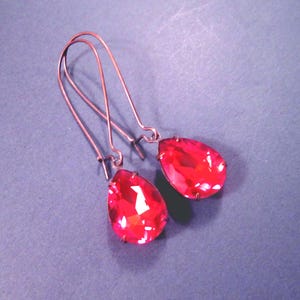 May include: A pair of long, copper-colored earrings with teardrop-shaped, faceted red crystal pendants. The earrings have a simple hook design and are displayed against a purple-gray background.
