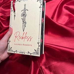 May include: A white book with a black border and a floral design. The title of the book is "Reckless" by Lauren Roberts. The book has a sword with a flower growing from it on the cover.