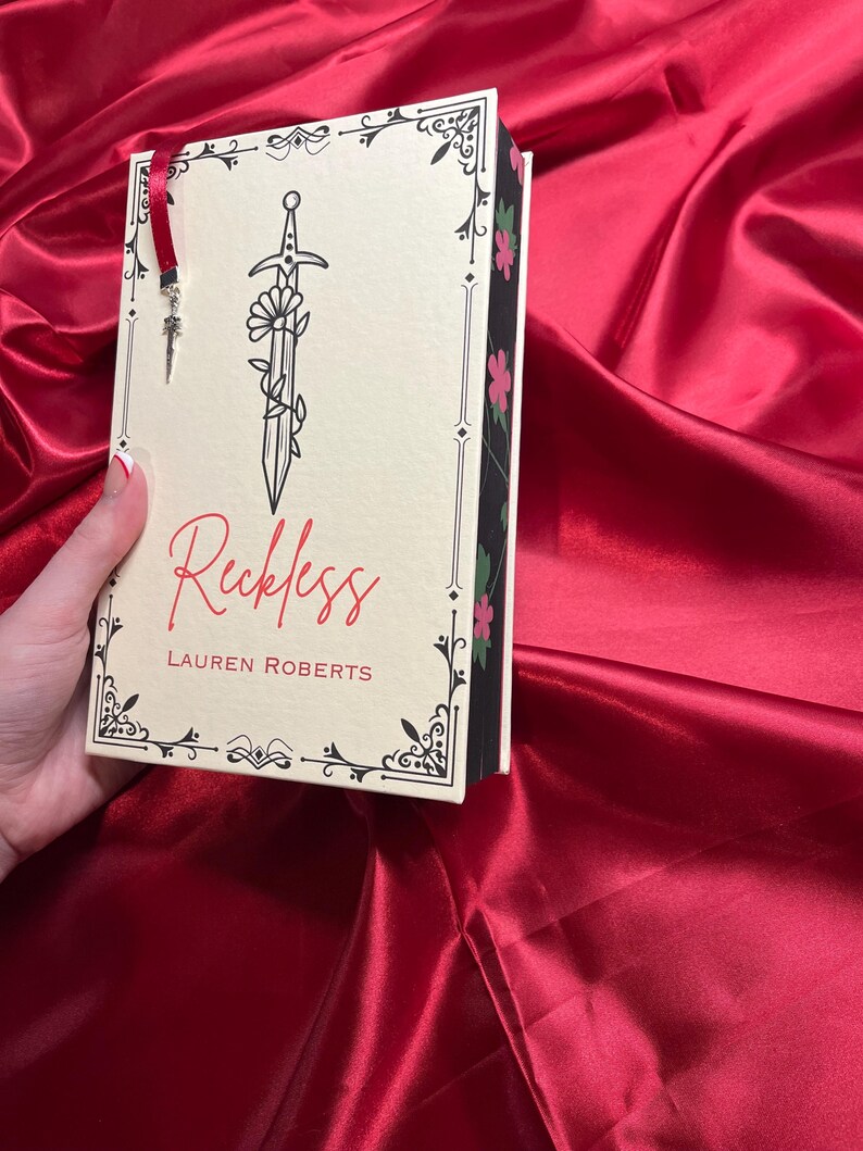 May include: A white book with a black border and a floral design. The title of the book is "Reckless" by Lauren Roberts. The book has a sword with a flower growing from it on the cover.