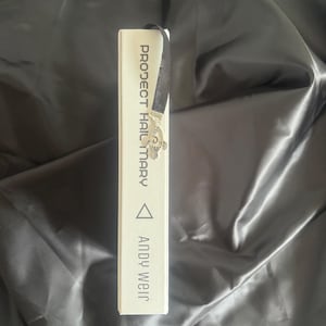 May include: A white book titled "PROJECT HAIL MARY" by Andy Weir. A black ribbon bookmark with a silver butterfly charm is inserted into the book. The book is on a black, satin-like surface.