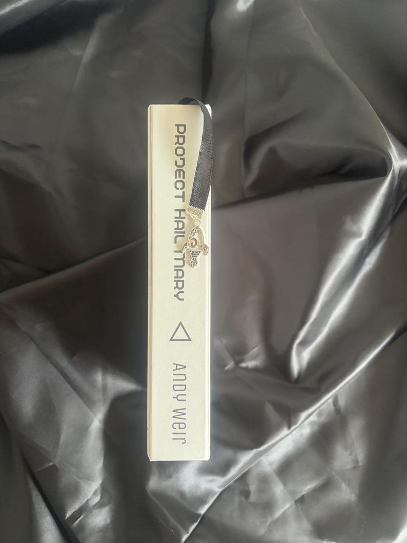 May include: A white book titled "PROJECT HAIL MARY" by Andy Weir. A black ribbon bookmark with a silver butterfly charm is inserted into the book. The book is on a black, satin-like surface.