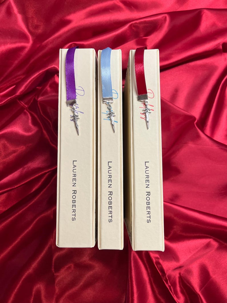 May include: Three hardcover books with matching ribbon bookmarks. Each bookmark has a silver charm in the shape of a sword. The books are all by the same author, Lauren Roberts.