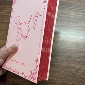 May include: A hardcover book titled "Rewind It Back" by Liz Tomforde. The book has a light pink cover with pink decorative accents and text. The spine is red with a repeating pattern of a boombox, hockey sticks, and the number 38.