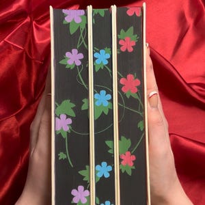 May include: Three hardcover books with black covers and a floral design. The flowers are purple, blue, and red.