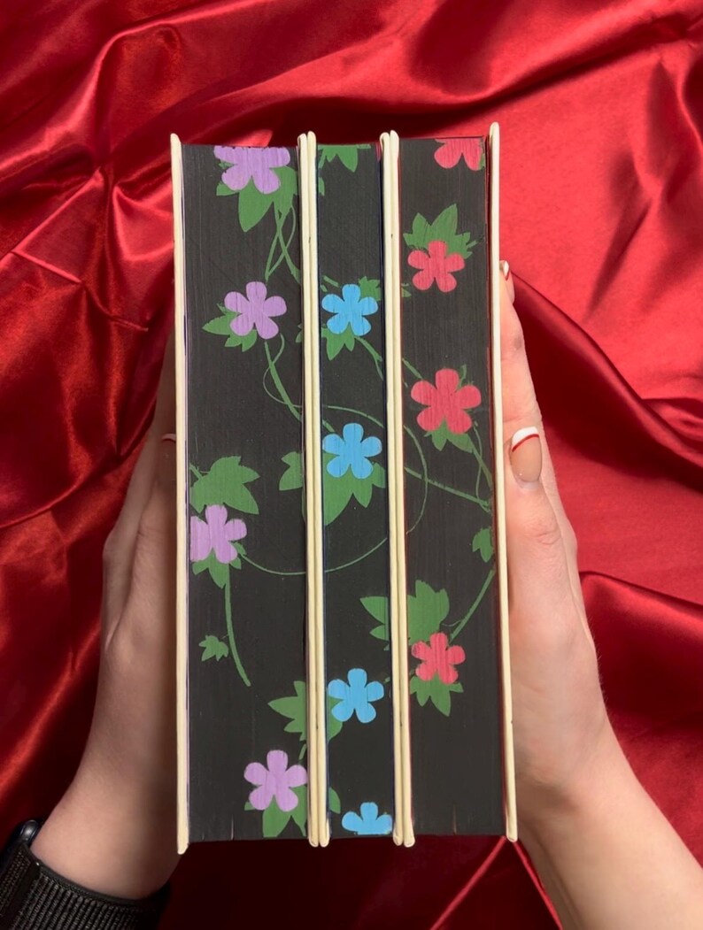 May include: Three hardcover books with black covers and a floral design. The flowers are purple, blue, and red.