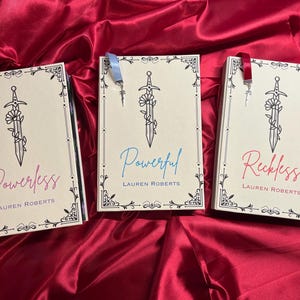 May include: Three paperback books with decorative borders and a sword design. The books are titled "Powerless", "Powerful", and "Reckless" with the author "Lauren Roberts" printed below each title. Each book has a ribbon bookmark.