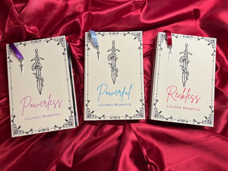 May include: Three paperback books with decorative borders and a sword design. The books are titled "Powerless", "Powerful", and "Reckless" with the author "Lauren Roberts" printed below each title. Each book has a ribbon bookmark.