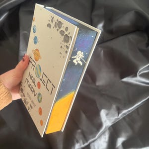 May include: A hardcover book titled "Project Hail Mary" by Andy Weir. The cover features planets and the title in white. The book's edge has a space-themed design with an astronaut. The book is held against a dark, silky background.
