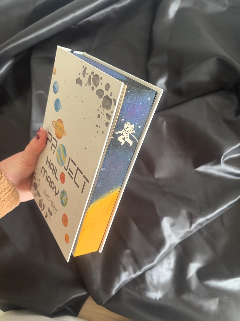 May include: A hardcover book titled "Project Hail Mary" by Andy Weir. The cover features planets and the title in white. The book's edge has a space-themed design with an astronaut. The book is held against a dark, silky background.