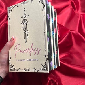May include: A stack of five books with floral patterned spines. The top book has a cream cover with a black and white illustration of a sword with a flower growing from it. The title "Powerless" is written in purple script and the author "LAUREN ROBERTS" is written in black below the title.