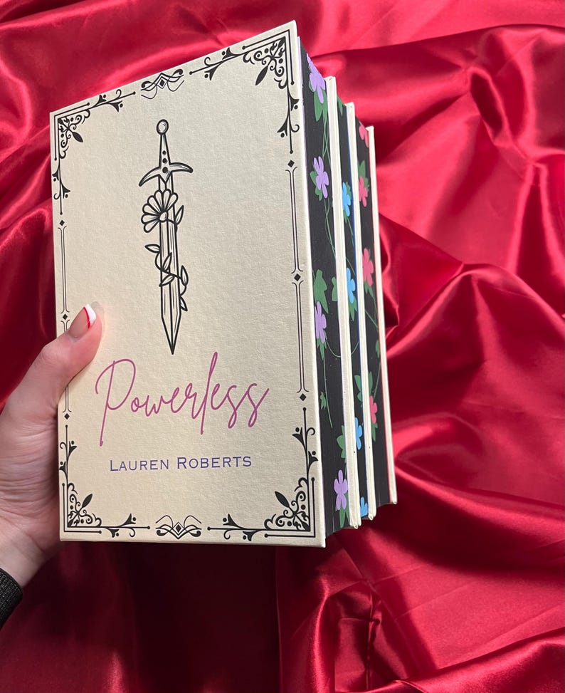 May include: A stack of five books with floral patterned spines. The top book has a cream cover with a black and white illustration of a sword with a flower growing from it. The title "Powerless" is written in purple script and the author "LAUREN ROBERTS" is written in black below the title.