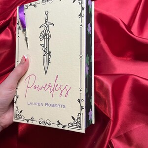 May include: A book titled "Powerless" by Lauren Roberts with a black and white illustration of a sword with flowers on the cover. The book has a purple and black spine with a floral design.