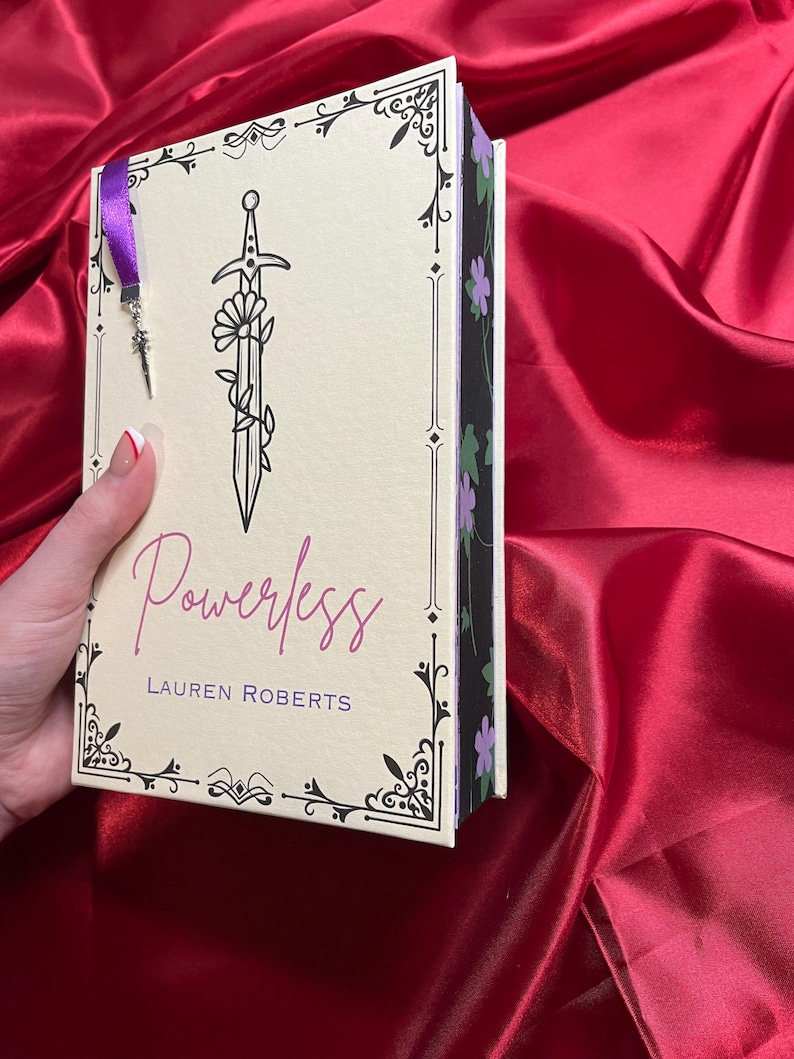 May include: A book titled "Powerless" by Lauren Roberts with a black and white illustration of a sword with flowers on the cover. The book has a purple and black spine with a floral design.