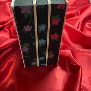 May include: Three hardcover books with black covers and floral designs. The books have white spines with a light blue, pink, and red floral pattern.