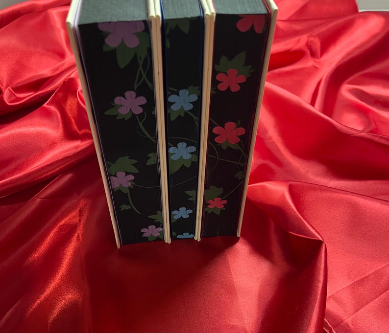 May include: Three hardcover books with black covers and floral designs. The books have white spines with a light blue, pink, and red floral pattern.