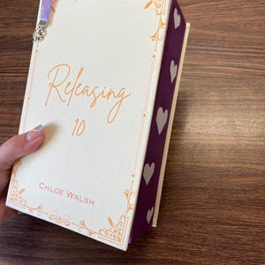 May include: A hardcover book with a cream-colored cover featuring the title "Releasing 10" in orange script, and the author's name, Chloe Walsh. The book has a purple spine with white heart shapes and a lavender ribbon bookmark with a silver charm.