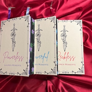 May include: Three paperback books with ornate borders and a sword and flower design. The titles are "Powerless", "Powerful", and "Reckless" with the author's name "Lauren Roberts" on each book.