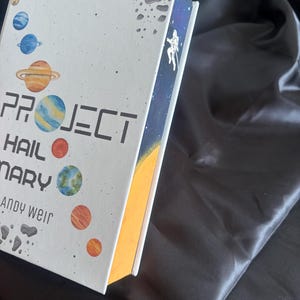 May include: A hardcover book titled "PROJECT HAIL MARY" by Andy Weir. The cover features a white background with colorful planets and asteroid illustrations. The spine shows an astronaut in space against a starry backdrop. The book's edges are painted yellow.