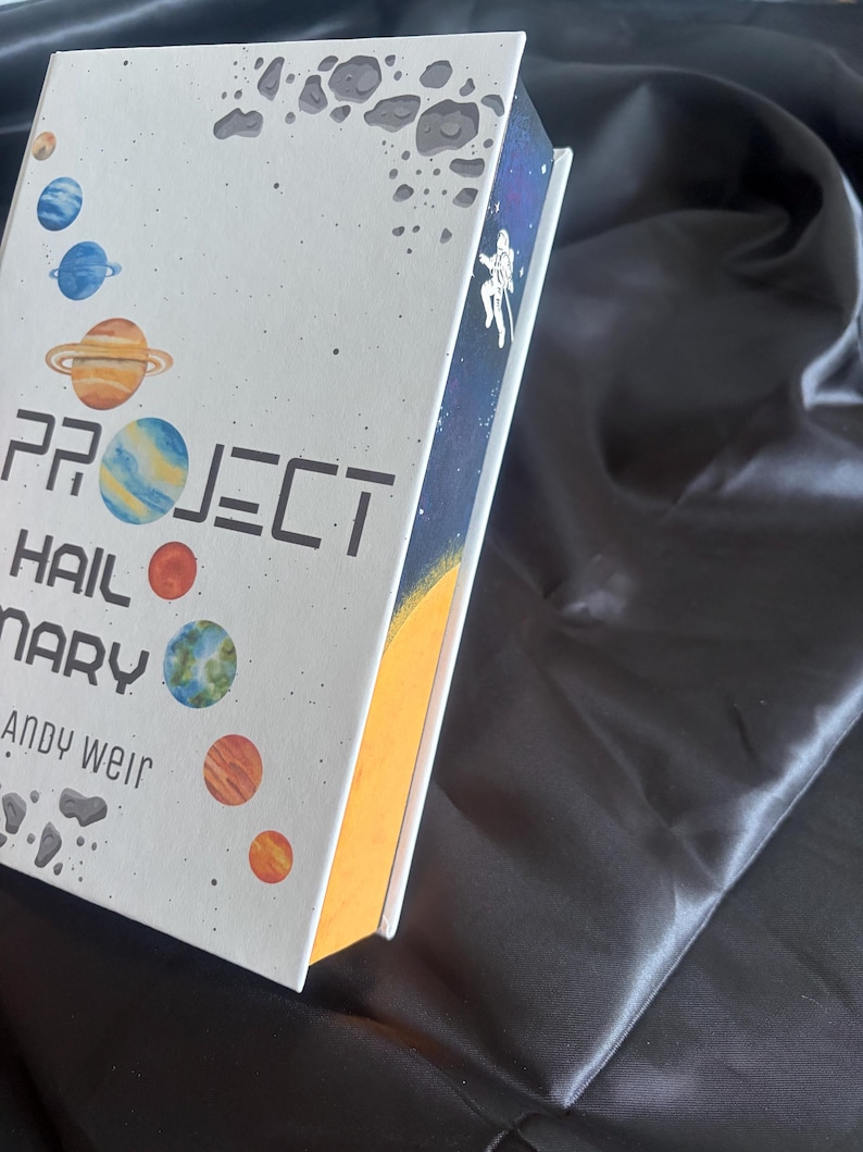 May include: A hardcover book titled "PROJECT HAIL MARY" by Andy Weir. The cover features a white background with colorful planets and asteroid illustrations. The spine shows an astronaut in space against a starry backdrop. The book's edges are painted yellow.
