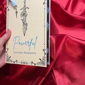 May include: A book titled "Powerful" by Lauren Roberts with a blue ribbon bookmark featuring a silver sword charm. The book cover has a black and white illustration of a sword with a flower.