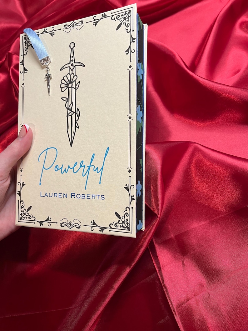 May include: A book titled "Powerful" by Lauren Roberts with a blue ribbon bookmark featuring a silver sword charm. The book cover has a black and white illustration of a sword with a flower.