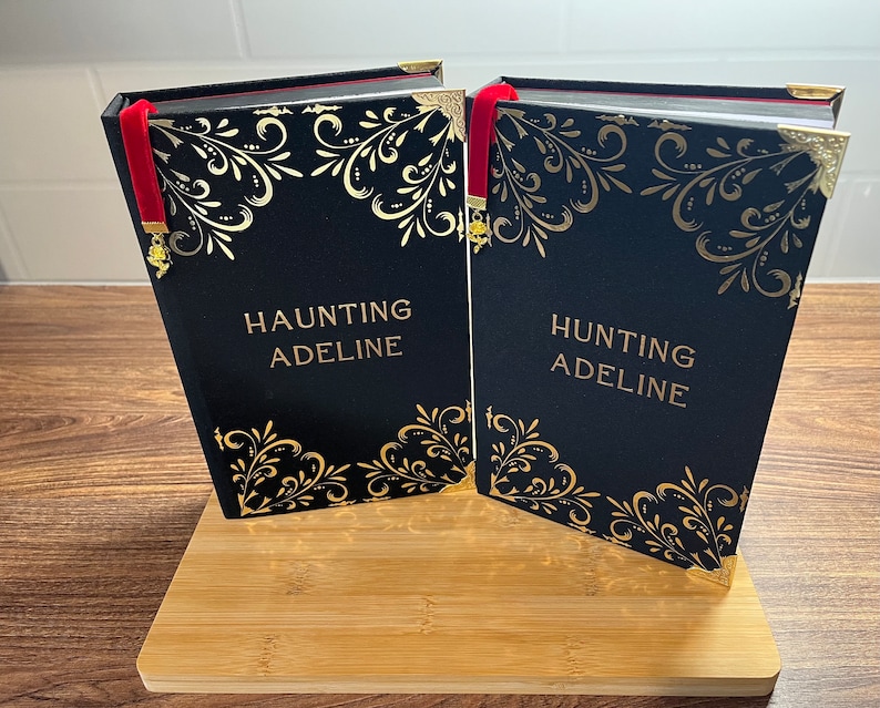 Może przedstawiać: Dwie czarne książki w twardej oprawie z złotymi akcentami i czerwonymi zakładkami. Książki noszą tytuły "Haunting Adeline" i "Hunting Adeline".