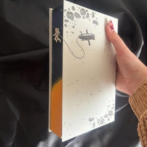 May include: A white book with an outer space theme. The cover features a space station and an astronaut tethered to it. The spine is dark blue with an astronaut and a yellow sun. Gray asteroids and stars are scattered across the cover.