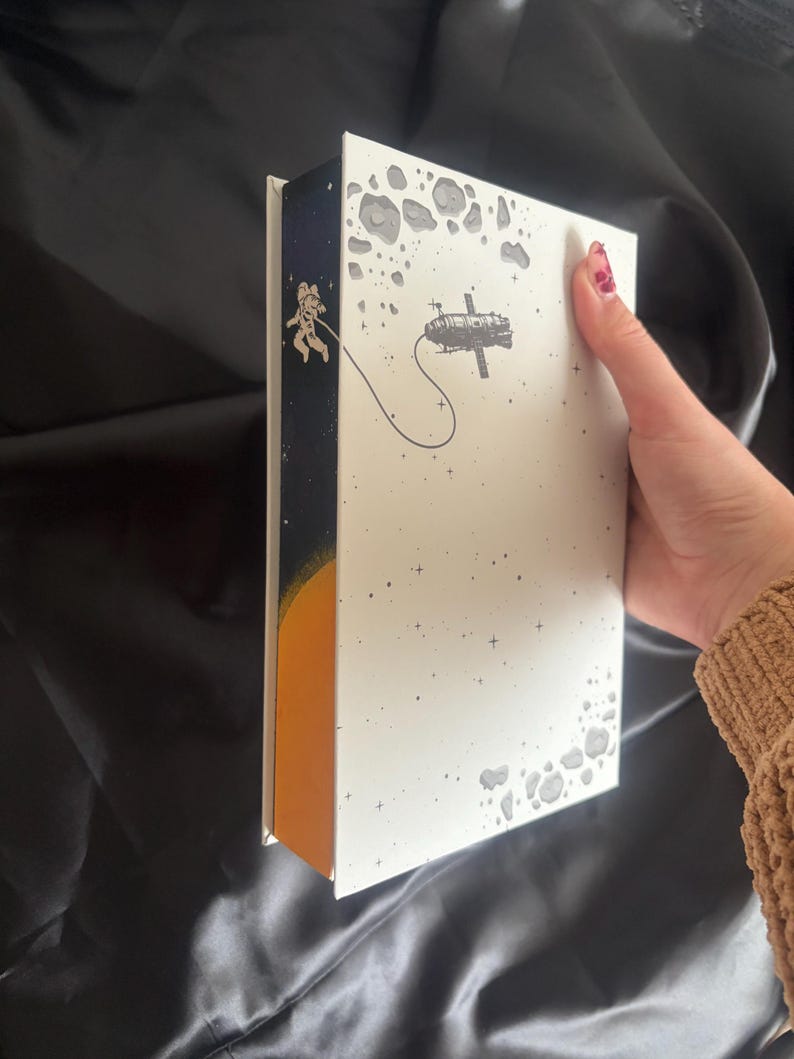 May include: A white book with an outer space theme. The cover features a space station and an astronaut tethered to it. The spine is dark blue with an astronaut and a yellow sun. Gray asteroids and stars are scattered across the cover.
