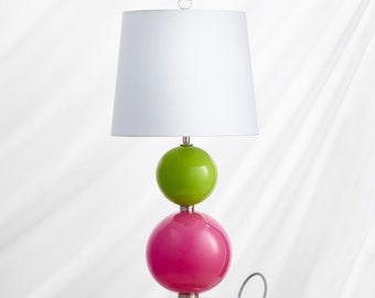 May include: A table lamp with a white lampshade and a base made of two stacked glass spheres. The top sphere is green and the bottom sphere is pink.