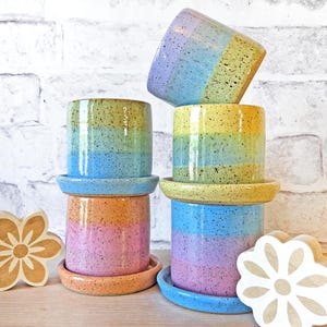 May include: A stack of six ceramic plant pots with matching saucers. The pots are cylindrical and feature a speckled, rainbow-colored glaze in shades of blue, yellow, orange, and pink. The pots are stacked on top of each other, with some tilted.