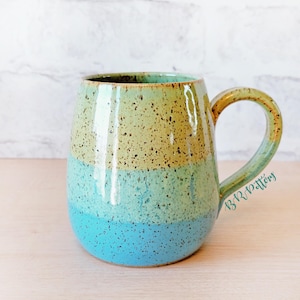 May include: A handmade ceramic mug with a speckled glaze. The mug features horizontal bands of color: blue at the base, transitioning to turquoise, and then to a tan-yellow at the top. The handle is a matching tan-yellow color.
