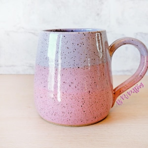 May include: A handmade ceramic mug with a speckled pink and lavender gradient. The mug has a rounded shape with a large handle and a glossy finish. The mug is suitable for hot or cold beverages.