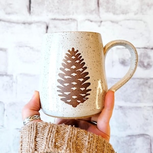 May include: A speckled ceramic mug with a large brown pinecone design. The mug has a curved handle and a neutral color palette, perfect for enjoying hot beverages. The mug is held in a person's hand.