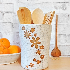 May include: A speckled white ceramic utensil holder with brown floral designs. The holder contains wooden spoons, spatulas, and a fork. A bowl of oranges sits to the left. A wooden spoon rests on the right.