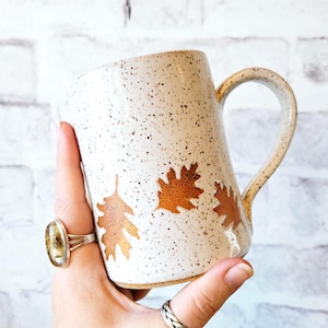 May include: A speckled white ceramic mug with three brown leaf designs. The mug has a curved handle and is held by a hand wearing silver rings. The mug is a neutral color and is suitable for hot or cold beverages.