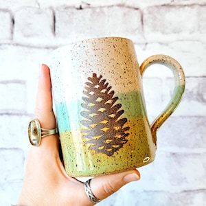 May include: A handmade ceramic mug with a pine cone design. The mug features a speckled cream top, transitioning to green and yellow, with a brown pine cone illustration. The handle and rim have a gold-like glaze. The mug is held in a hand.