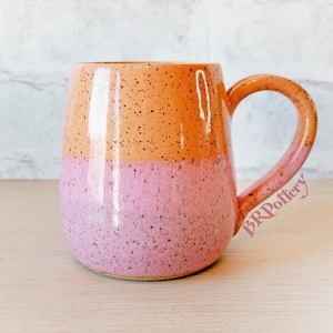 May include: A handmade ceramic mug with a two-tone design. The top half is orange with black speckles, and the bottom half is pink with black speckles. The mug has a large, rounded handle and the text "BRPottery" is visible on the side.