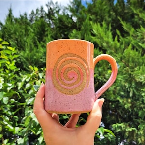 May include: A handmade ceramic mug with a spiral design. The mug features a gradient of colors, transitioning from orange at the top to pink at the bottom. The spiral is a contrasting tan color. The mug has a curved handle.