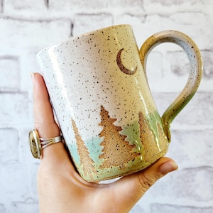 May include: A ceramic mug with a nature-inspired design. The mug features a speckled white background with a brown crescent moon and brown and green trees. The handle and base have a green and brown glaze.