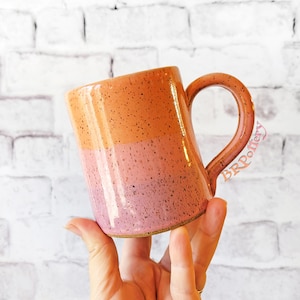 May include: A two-tone ceramic mug with a speckled finish. The top section is orange, fading into pink below. The mug has a large handle and is held in a hand. The mug is likely handmade.
