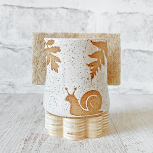 May include: A ceramic sponge holder with a speckled white glaze, featuring a brown snail and leaf pattern. The holder sits on a light-colored, fluted wooden base and contains a rectangular, tan-colored fibrous scrubbing sponge.