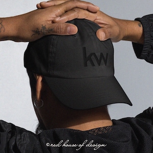 May include: A black baseball cap featuring the letters "kw" embroidered on the front. The cap has a curved peak and is made from a soft fabric. The image also includes the text "red house of design".