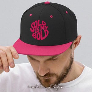 May include: A black and pink flat-peak cap featuring the text "SOLD IS MY BOLD" in pink. The cap has a pink peak and a pink button on top. The text is in a bold, rounded font.