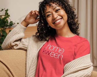 May include: A coral-colored t-shirt with the text "home kween" in white and pink lettering. The shirt is worn with a beige cardigan. The person is smiling.