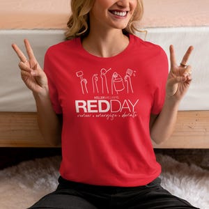 Puede incluir: Una camiseta roja con un diseño gráfico blanco que representa un grupo de manos sosteniendo herramientas y un cubo. El texto "Keller Williams REDDAY renew. energize. donate." está impreso debajo del gráfico.