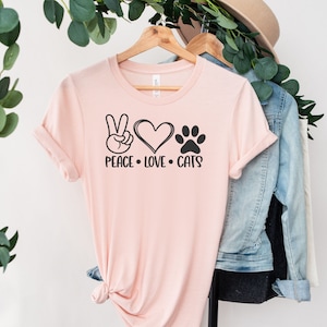 May include: A pink t-shirt with a peace sign, heart, and paw print graphic. The text on the shirt reads "Peace Love Cats".