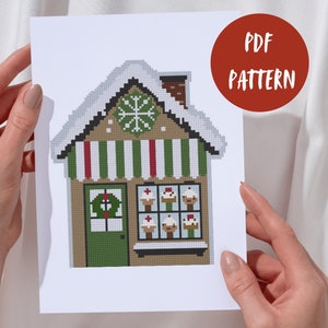 Puede incluir: Un patrón PDF imprimible para bordar a punto de cruz una casa de jengibre con un toldo verde, rayas rojas y blancas, una corona en la puerta y una ventana llena de cupcakes. La casa está cubierta de nieve blanca.