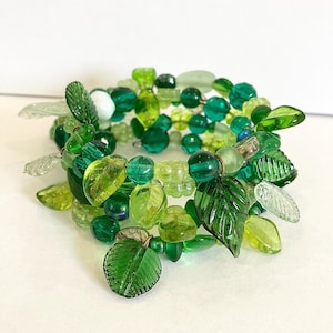 Könnte beinhalten: Ein grünes Glasperlenarmband mit blattförmigen Anhängern. Das Armband besteht aus mehreren Perlensträngen in verschiedenen Grüntönen, darunter Hellgrün, Dunkelgrün und Smaragdgrün. Die blattförmigen Anhänger sind ebenfalls aus Glas und haben eine strukturierte Oberfläche.